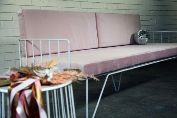 BLUSH PINK COUCH 05_resized Blush pink velvet couch with sleek metal legs set against a white brick wall, ideal for events and available for hire in Perth.