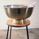 A large golden metal bowl resting on a wooden stand, serving as an elegant drinks bucket for event hire.